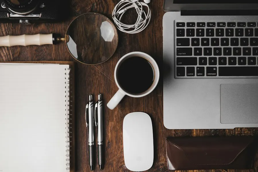 Overhead view of a wooden desk with a laptop, a cup of coffee, a spiral notebook, two pens, a mouse and a magnifying glass