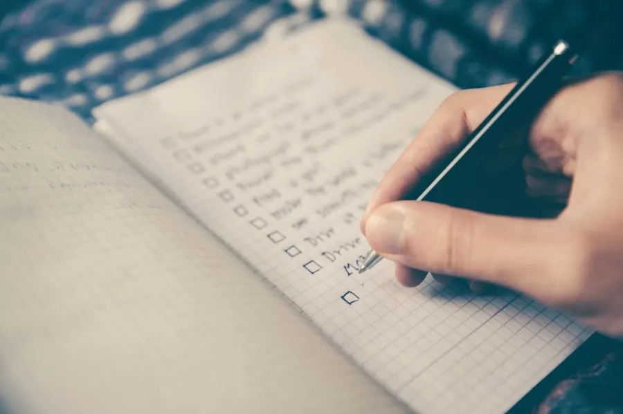 A hand holding a pen writes a checklist in a grid-paper notebook, with an empty checkbox beside each item