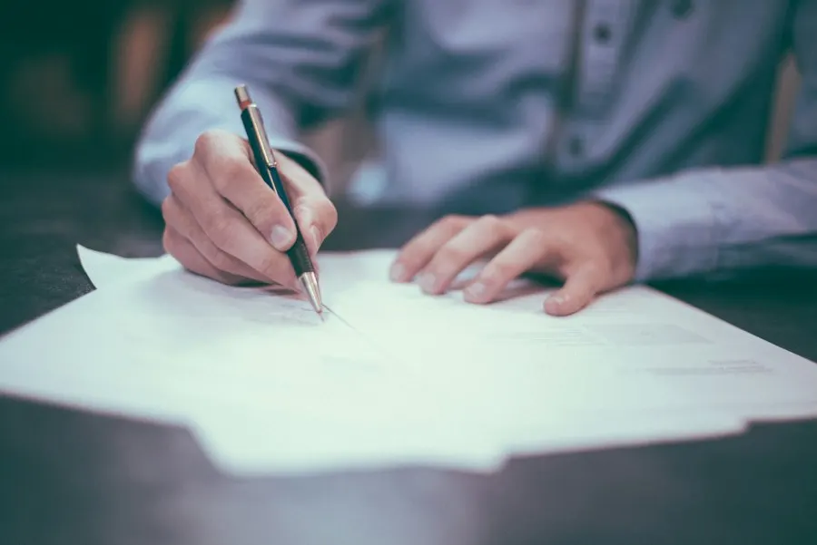 A person in a blue shirt writes on a sheet of paper with a pen at a dark table