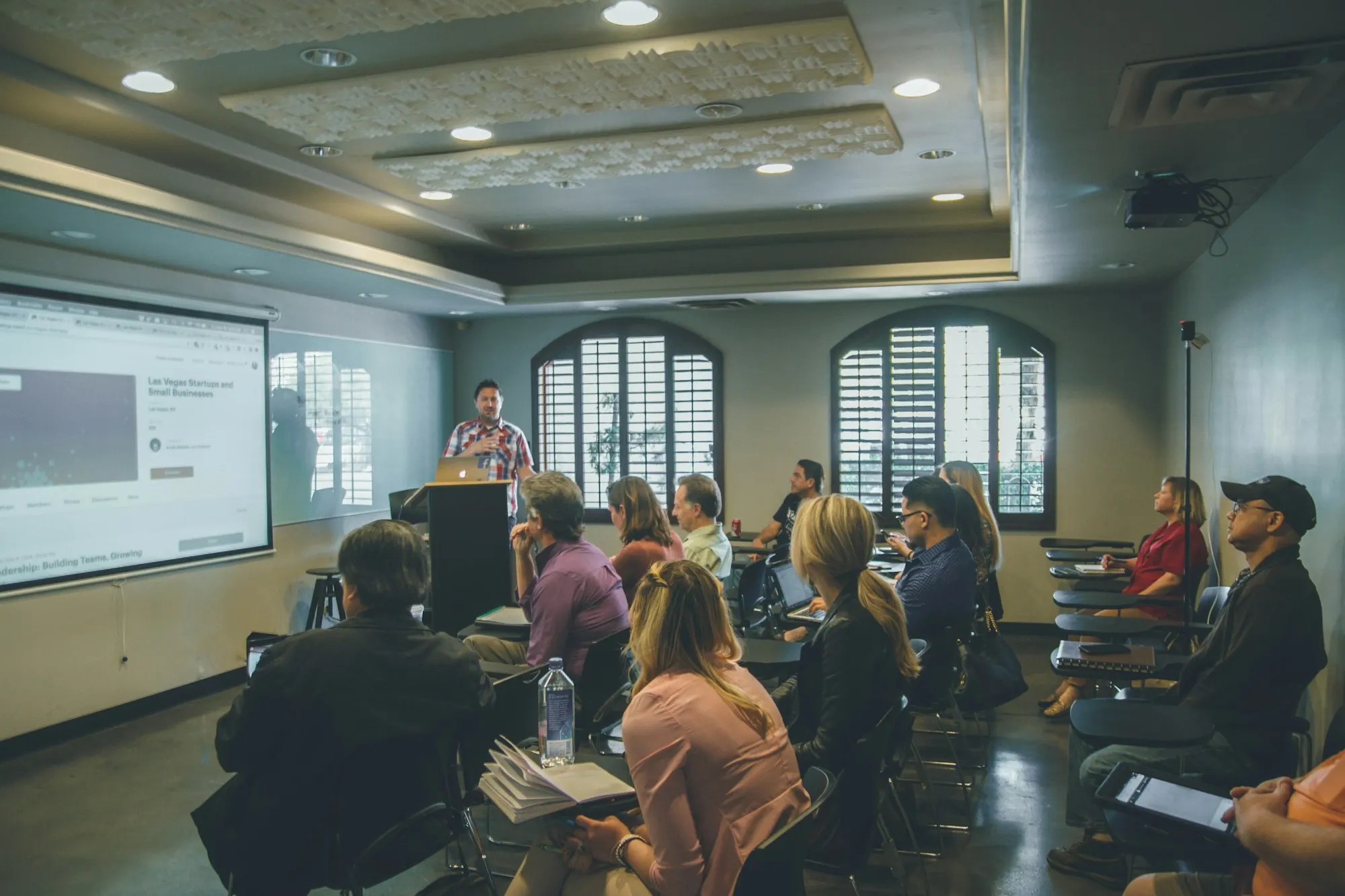 Adults seated in rows in a bright meeting room listen to a presenter standing beside a large projected screen
