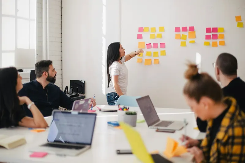 A woman points to rows of sticky notes on a wall while a group with laptops listens around a white table