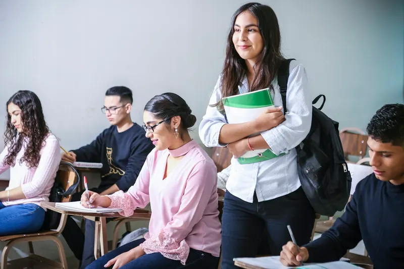 A smiling young woman with a backpack and a binder stands among young adults taking notes at classroom desks