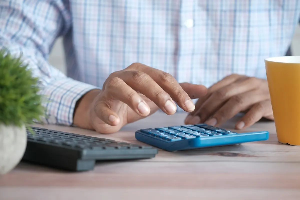 A person in a checked shirt works a blue calculator on a desk beside a keyboard, a small plant and a yellow mug
